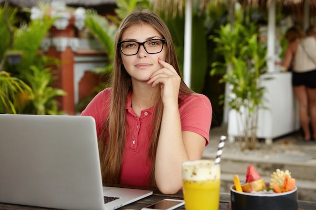 Atraente mulher autônoma de óculos trabalhando no projeto, usando wi-fi em seu laptop, sentada à mesa de madeira com bebida gelada e cópia espaço celular, com expressão séria