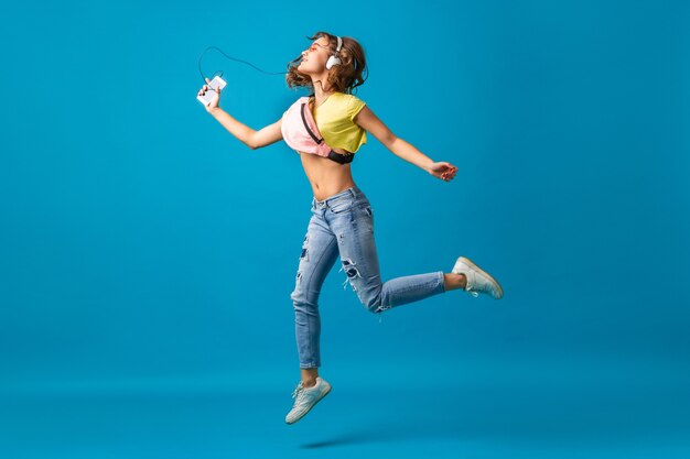 Atraente mulher sorridente feliz dançando ouvindo música em fones de ouvido, vestida com roupa elegante hipster isolada no fundo azul do estúdio, usando roupas coloridas