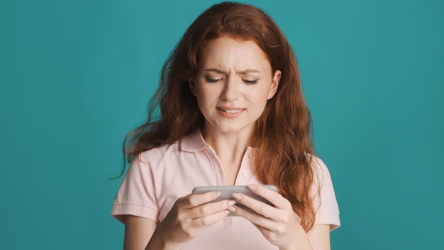 Atraente ruiva nervosa jogando no jogo no smartphone sobre fundo colorido expressão chateada