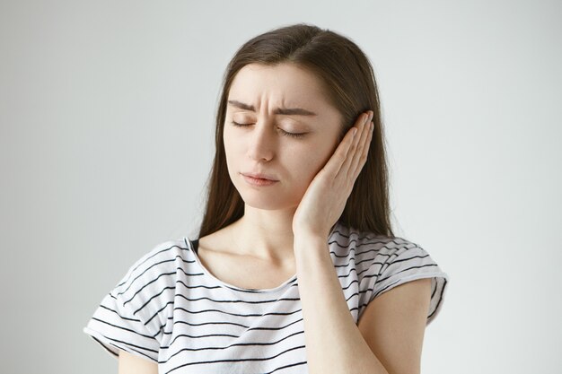 atraente, vestida casualmente, jovem, morena, mulher, mantendo os olhos fechados e cobrindo a orelha dolorida com a mão, embora tenha alguns problemas de audição. Conceito de saúde, pessoas e dor