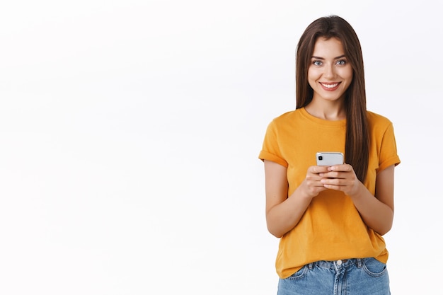 Foto grátis atrevida e feliz mulher bonita em uma camiseta amarela, segurando uma câmera de aparência de smartphone satisfeita e alegre, fazendo compras online, baixe o aplicativo móvel para editar a foto e postar na rede social