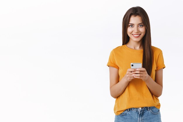 Atrevida e feliz mulher bonita em uma camiseta amarela, segurando uma câmera de aparência de smartphone satisfeita e alegre, fazendo compras online, baixe o aplicativo móvel para editar a foto e postar na rede social
