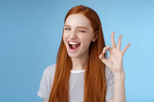 Atrevida garota ruiva atraente piscando misteriosamente sorrindo amplamente dar sinal de aprovação mostrar ok ok excelente gesto satisfeito boa escolha concordar ótima decisão, em pé fundo azul encantado.