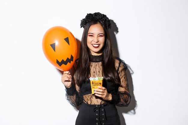 Atrevida linda mulher asiática desfrutando de doces ou travessuras, comemorando o dia das bruxas, segurando um balão laranja e doces.