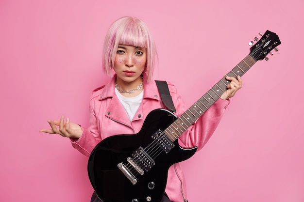 Auto-confiante roqueira na moda com cabelo rosado se prepara para o festival de música pratica violão usa jaqueta poses contra a parede rosa. músico talentoso toca instrumento musical