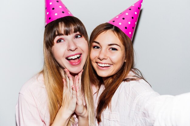 Auto-retrato de duas mulheres sorridentes com chapéus de aniversário de papel rosa. Amigos de pijama rosa.