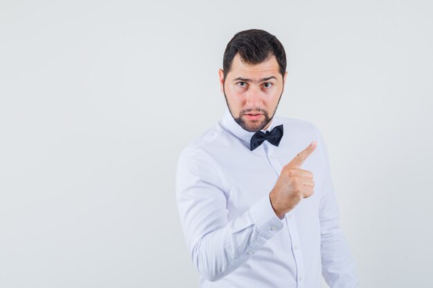 Aviso masculino jovem com o dedo na camisa branca e olhando sério, vista frontal.