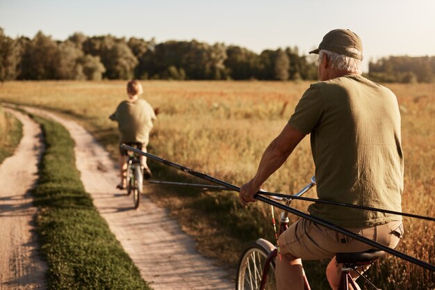 Avô e seu neto vão pescar em bicicletas, vista traseira da família no Prado em bicicletas com varas de pesca, homem sênior e jovem vestindo fechamento casual, belo campo e árvores.