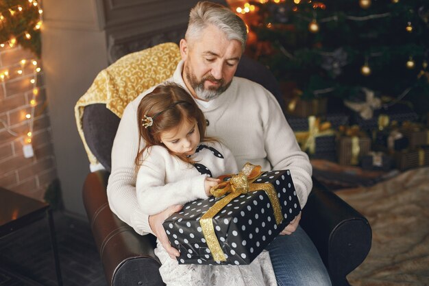 Avô sentado com sua neta. Comemorando o Natal em uma casa aconchegante. Homem com uma camisola de malha branca.