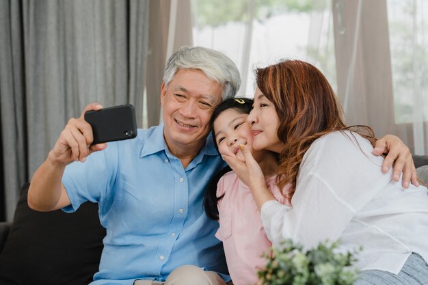 Avós asiáticos selfie com neta em casa. O chinês sênior, o vovô e a avó felizes passam o tempo da família relaxam usando o telefone celular com a criança da moça que encontra-se no sofá no conceito da sala de visitas.