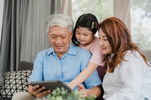 Avós e neta asiáticos usando o tablet em casa. O chinês sênior, o vovô e a avó felizes passam o tempo em família relaxam com a menina que verifica as mídias sociais, deitado no sofá no conceito da sala de estar