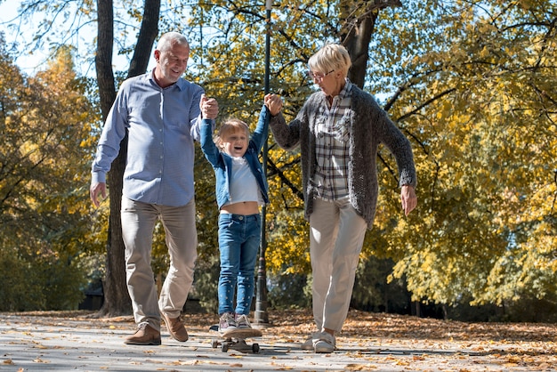 Avós sorridentes de mãos dadas com a neta enquanto ela anda de skate em um parque de outono