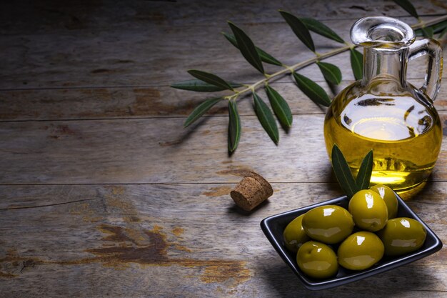 Azeitonas de aparência saborosa, azeite de oliva extra virgem e folhas de oliveira em fundo escuro de madeira