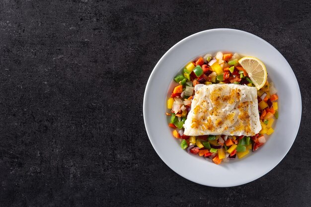 Bacalhau grelhado com legumes no prato sobre fundo preto de pedra