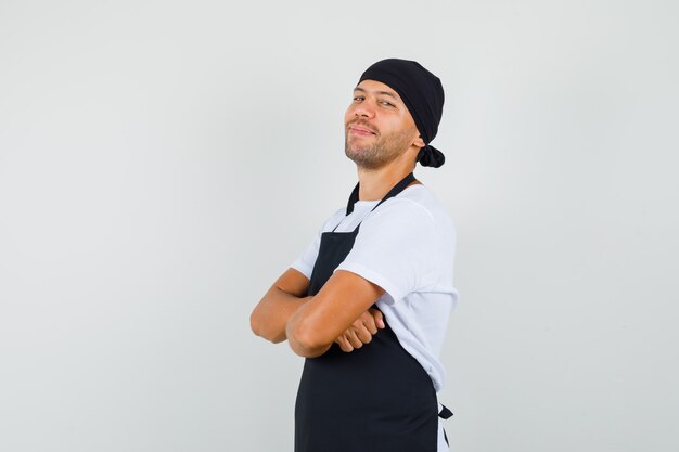 Baker man em t-shirt, avental em pé com os braços cruzados e olhando alegre