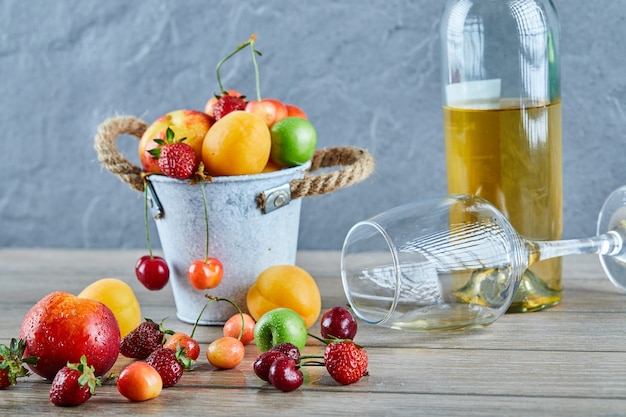 Balde de frutas frescas de verão, garrafa de vinho branco e copo vazio na mesa de madeira.