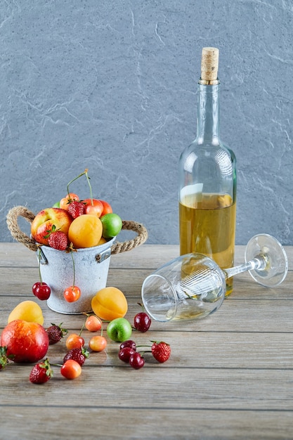 Balde de frutas frescas de verão, garrafa de vinho branco e copo vazio na mesa de madeira.