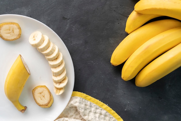 Banana fresca fatiada em um prato de cerâmica branca na mesa preta, preparando ingredientes para um café da manhã saudável