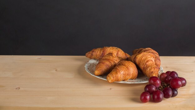 Bando de uvas vermelhas e placa de croissant na mesa contra o fundo preto