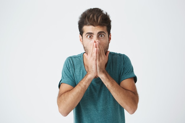 Barba por fazer atraente jovem hispânico em camiseta azul, cobrindo o rosto com a mão, olhando com total descrença depois de receber más notícias.