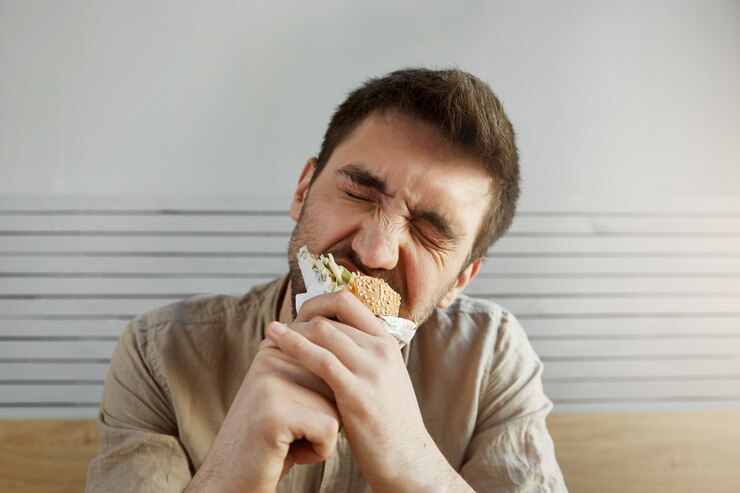 Foto grátis barba por fazer bonito jovem com cabelos escuros, comendo sanduíche em fast-food com os olhos fechados, com expressão feliz e satisfeita.