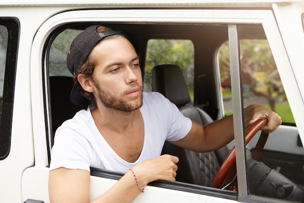 Barba por fazer jovem bonito vestindo camiseta casual e boné de beisebol para trás, olhando pela janela aberta de seu veículo com tração nas quatro rodas, indo em viagem de safari