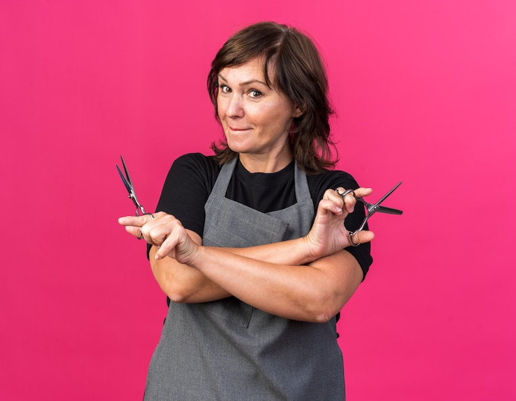 Foto grátis barbearia adulta, branca, feminina, de uniforme, segurando uma tesoura e olhando para a câmera, isolada em um fundo rosa com espaço de cópia
