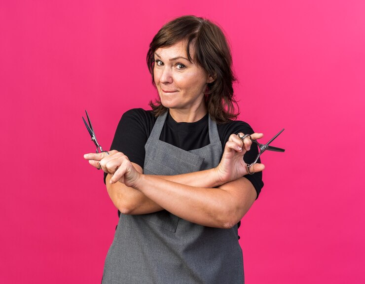 Foto grátis barbearia adulta, branca, feminina, de uniforme, segurando uma tesoura e olhando para a câmera, isolada em um fundo rosa com espaço de cópia