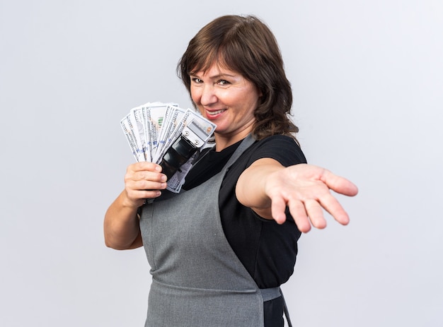 Barbeira adulta, branca, feminina, sorridente, de uniforme, segurando o aparador de cabelo com dinheiro e estendendo a mão, isolada na parede branca com espaço de cópia