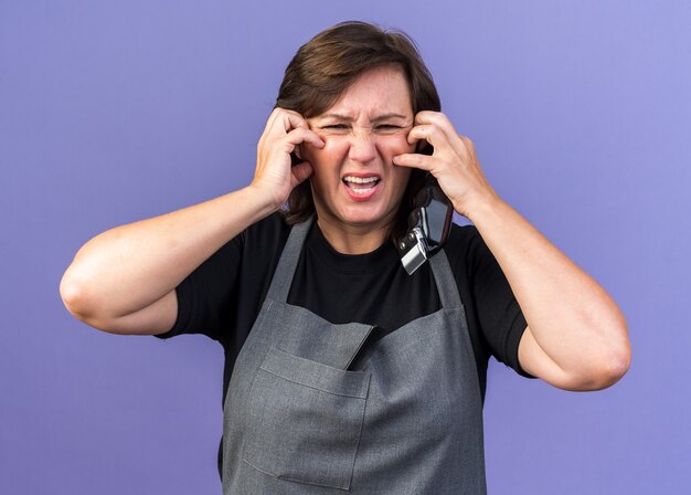 Barbeira adulta, caucasiana, adulta, de uniforme, coçando o rosto com as mãos segurando uma tesoura de cabelo isolada na parede roxa com espaço de cópia