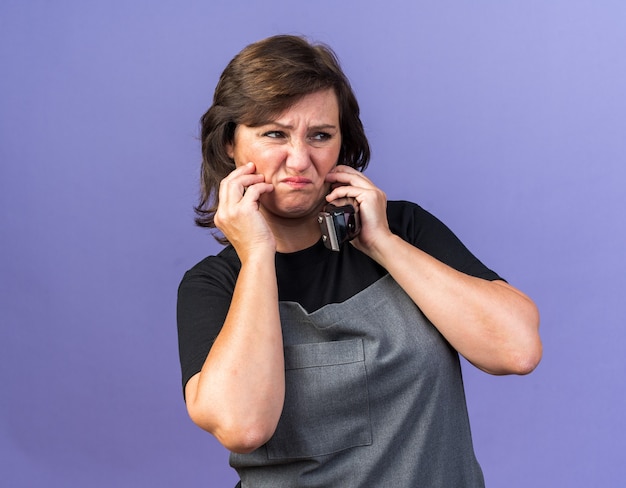 Barbeira adulta, caucasiana, feminina, adulta, de uniforme, colocando as mãos no rosto, segurando o aparador de cabelo e olhando para o lado isolado no fundo roxo com espaço de cópia