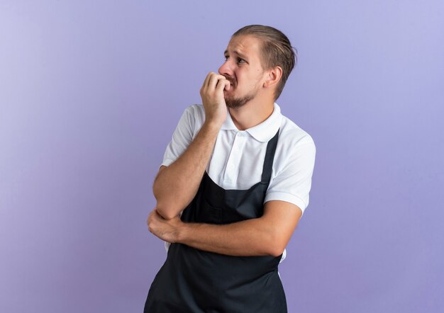 Barbeiro bonito jovem ansioso vestindo uniforme olhando para o lado, colocando os dedos nos lábios e a mão sob o cotovelo isolado em roxo com espaço de cópia