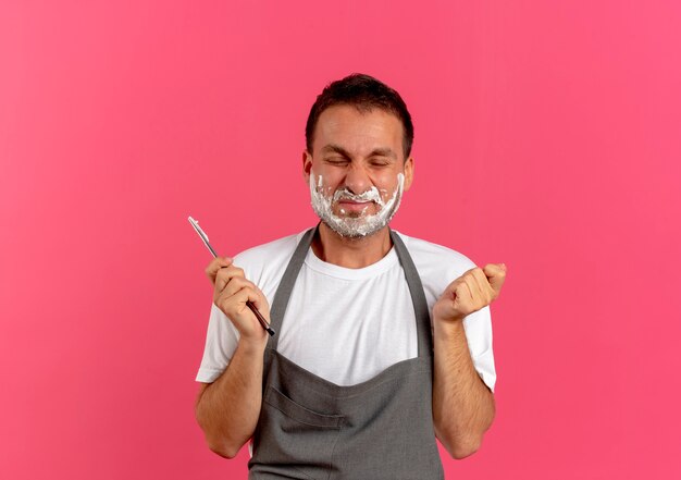 Barbeiro de avental com espuma de barbear no rosto segurando uma navalha, parecendo muito infeliz com os olhos fechados em pé sobre uma parede rosa