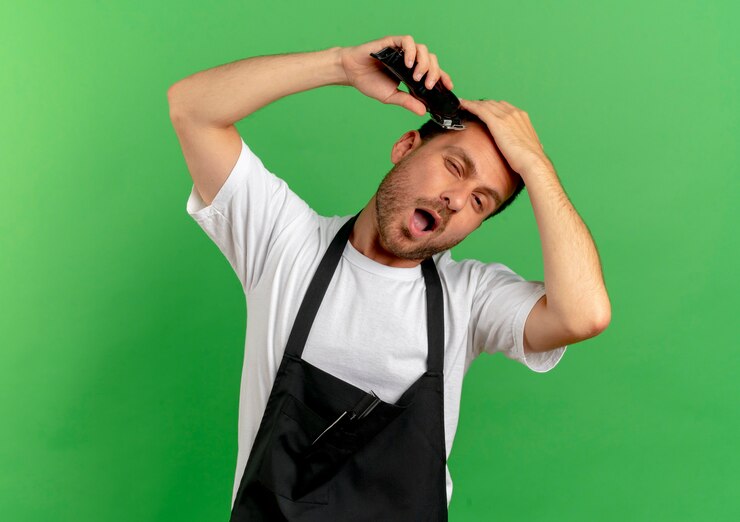 Foto grátis barbeiro de avental raspando o cabelo com uma máquina de corte de cabelo parecendo confiante em pé sobre uma parede verde