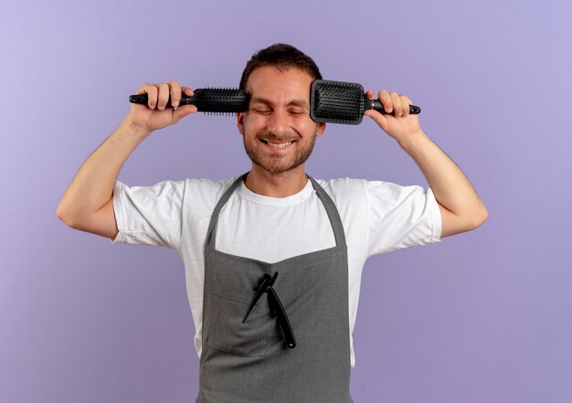 Foto grátis barbeiro de avental segurando escovas de cabelo e sorrindo alegremente com os olhos fechados em pé sobre uma parede roxa