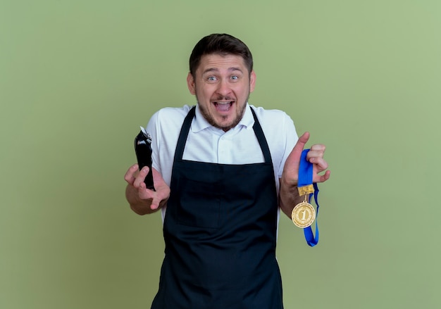 Barbeiro de avental segurando um aparador de barba e medalha de ouro olhando para a câmera animado e feliz em pé sobre o fundo verde