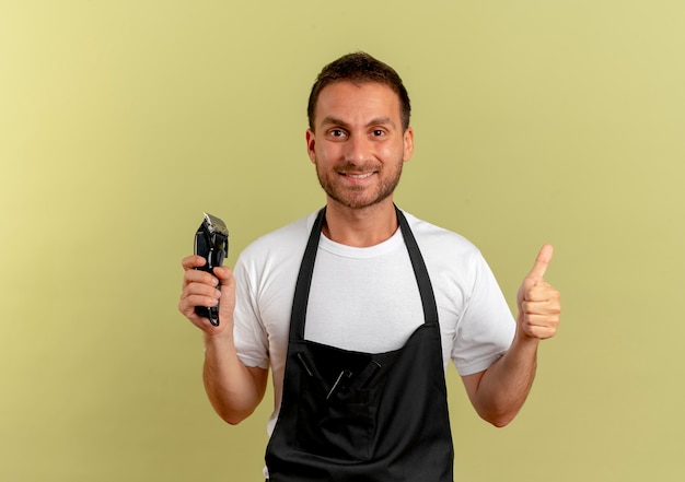 Foto grátis barbeiro de avental segurando um aparador e sorrindo alegremente mostrando os polegares em pé sobre a parede de luz