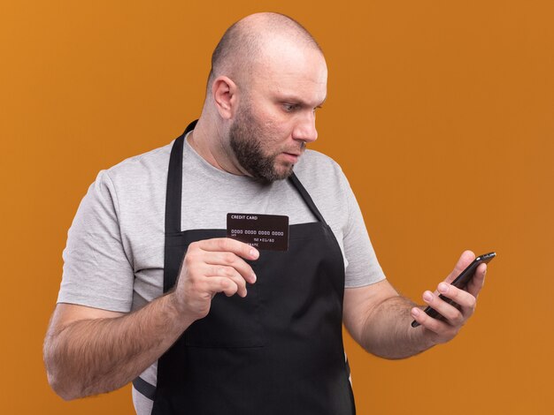 Barbeiro de meia-idade confuso, de uniforme, segurando um cartão de crédito e olhando para o telefone na mão, isolado na parede laranja