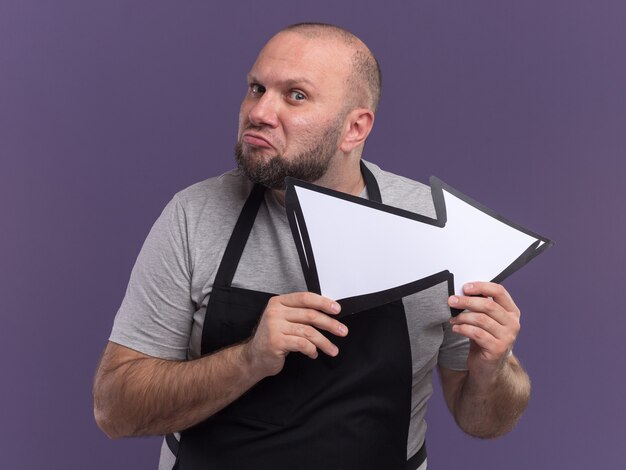 Barbeiro eslavo confuso, de meia-idade, de uniforme, segurando a marca de direção isolada na parede roxa