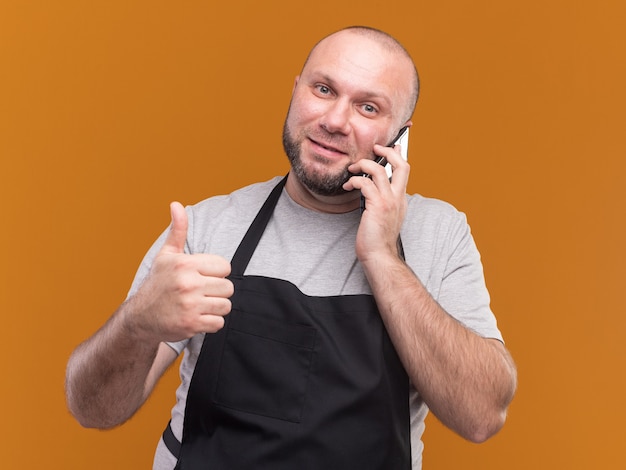 Barbeiro eslavo de meia-idade, de uniforme, satisfeito falando ao telefone, mostrando o polegar isolado na parede laranja