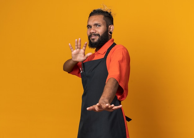 Barbeiro jovem afro-americano descontente, vestindo uniforme em pé na vista de perfil, fazendo gesto de recusa isolado na parede laranja com espaço de cópia