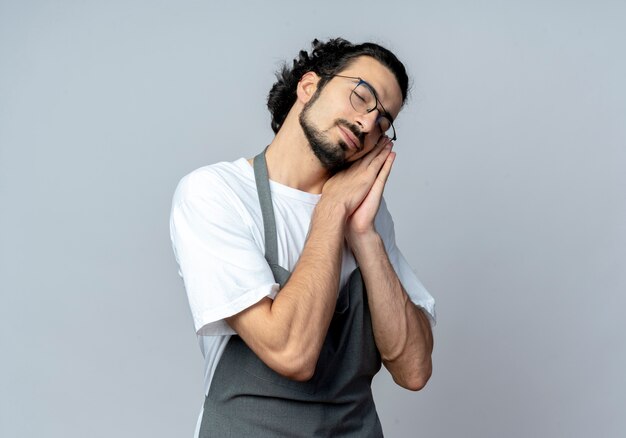 Barbeiro jovem caucasiano com sono, usando óculos e faixa de cabelo ondulado em uniforme, fazendo gestos para dormir com os olhos fechados, isolado no fundo branco com espaço de cópia