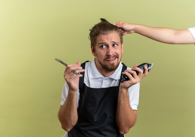 Barbeiro jovem e bonito confuso segurando uma tesoura e uma tesoura de cabelo olhando para o lado de alguém penteando seu cabelo isolado em verde oliva com espaço de cópia