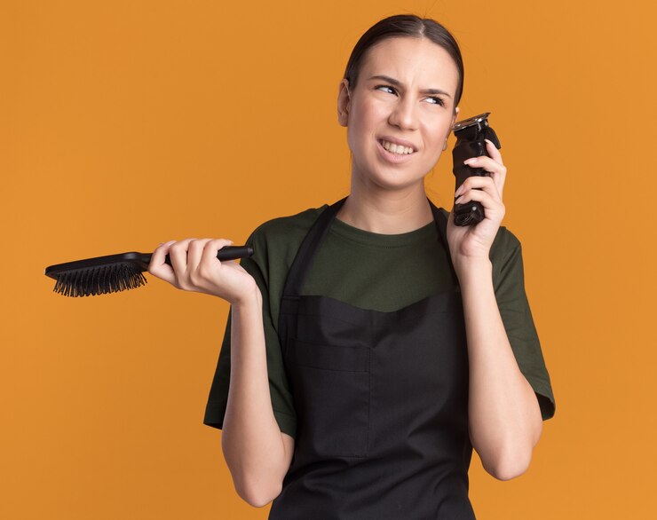 Foto grátis barbeiro jovem morena desagradável de uniforme segurando uma tesoura de cabelo e um pente olhando para o lado laranja