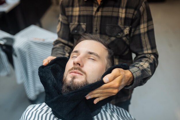 Barbeiro preparando o rosto do homem para fazer a barba com toalha quente no rosto na barbearia