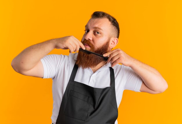 Barbeiro profissional barbudo com avental penteando a barba em pé sobre a parede laranja