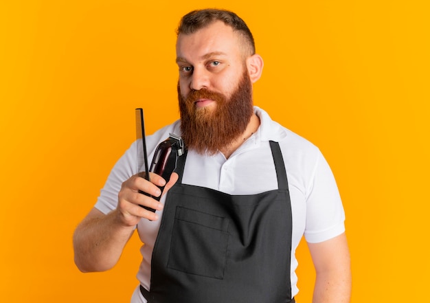 Barbeiro profissional barbudo com avental segurando uma máquina de barbear e uma escova de cabelo, parecendo confiante em pé sobre a parede laranja