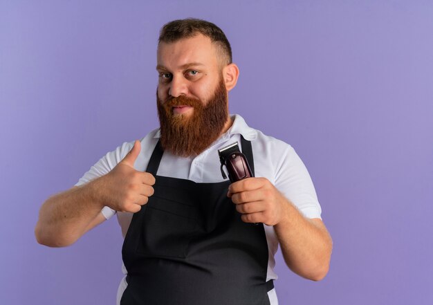 Barbeiro profissional barbudo com avental segurando uma máquina de corte de cabelo e sorrindo amigável mostrando os polegares em pé sobre a parede roxa