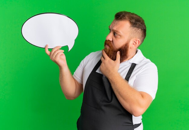 Barbeiro profissional barbudo de avental segurando um cartaz de balão de fala em branco, olhando para ele perplexo em pé sobre a parede verde