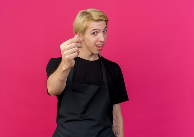 Barbeiro profissional com avental olhando para a frente sorrindo, fazendo gesto de dinheiro, esfregando os dedos em pé sobre a parede rosa